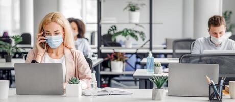 Working with client by phone remotely during COVID-19 outbreak. Millennial woman in protective mask talking at smartphone and typing on laptop in modern office interior, copy space photo