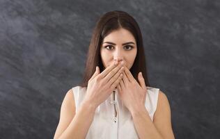 mantener silencio. asustado mujer cubierta boca con manos mientras posando a cámara en gris estudio antecedentes. conmocionado niña cerca labios con palmas, hablar No mal concepto foto