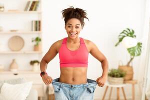Successful Weight Loss. Happy African Woman After Slimming Showing Her Old Oversize Pants Standing Indoors, Smiling To Camera photo