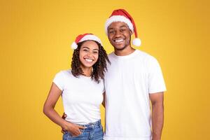 Christmas. Joyful African American Couple In Santa Hats Posing Smiling To Camera Standing Hugging On Yellow Studio Background. Merry Xmas And Happy New Year Advertisement, X-Mas Celebration Concept photo