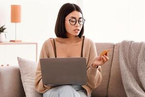 Online Banking And E-commerce Concept. Asian woman in glasses making purchases on internet, using laptop, holding credit card, looking at cvv, checking security code for safe transfer transaction photo