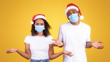 Christmas In Coronavirus Pandemic. Puzzled Black Couple Shrugging Shoulders Wearing Protective Face Masks And Xmas Santa Hat Standing On Yellow Studio Background. Holidays On Self-Isolation. Panorama photo