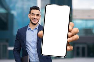 Handsome Arab Businessman In Suit Demonstrating Big Blank Smartphone At Camera, Excited Middle Eastern Male Entrepreneur Recommending New Mobile App, Posing Outdoors Near Business Center, Mockup photo