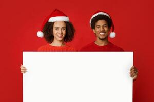 Christmas couple holds white board with copy space and advertise New Year season shopping sale concept. Millennial african american happy people in hats, isolated on red background, studio shot photo