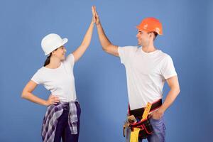 Happy couple in protective hard hats giving high five to each other while renovating their house, enjoying making flat repairment together, standing over blue studio background, free space photo