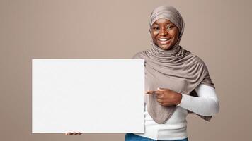 Happy african muslim woman in hijab pointing at blank white placard in her hands, showing free copy space for your design or advertisement, standing over light studio background, looking at camera photo