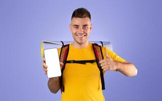 Online trip booking concept. Handsome young man showing mobile phone with empty screen and making thumb up gesture on blue studio background. Space for your website design photo