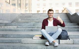 Happy student sitting on the stairs showing thumb up working on laptop, in the university campus. Technology, education and remote working concept, copy space photo