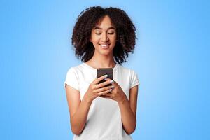 Texting message and modern blog. Smiling millennial african american lady in pink t-shirt with curly hair, holding smartphone, writing and looking at device, empty space, studio shot, cut out photo