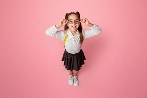 Success in school. Smiling little schoolgirl with glasses and backpack shows tongue and have fun, isolated on pink background, studio shoot photo