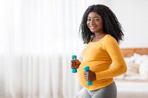 Active and sportive pregnancy concept. Happy pregnant black woman lifting dumbbells, exercising at home, free space. Cheerful and healthy african american expecting lady training in bedroom photo