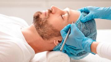 Handsome man getting pencil marks on forehead before cosmetic surgery or correction, ready for operation. Plastic surgeon applying marks on man face before surgery at aesthetic clinic, side view photo