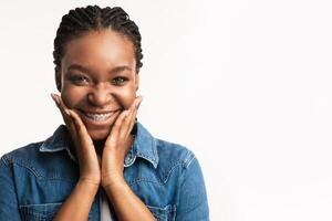 Orthodontics Concept. Joyful Black Girl With Brackets Cupping Face In Hands Smiling To Camera On White Background. Studio Shot photo