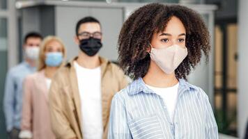 New normal and temperature check before work. Millennial multiracial employees in protective masks stand in line with social distancing at office in morning, free space, blurred, indoors photo