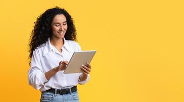 Young Brunette Woman Using Digital Tablet, Browsing Internet Or Social Networks, Standing Over Yellow Studio Background, Enjoying Modern Technologies, Checking New App Or Website, Copy Space photo