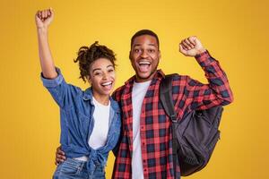 Joyful African American Couple With Backpack Shaking Fists, Excited About End Of Lockdown Posing Over Yellow Studio Background. Vacation, Finally photo