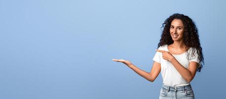 Positive Young Curly Woman Pointing At Something On Her Open Palm, Demonstrating Invisible Object, Recommending Something, Standing Over Purple Background, Free Place For Mockup, Panorama photo