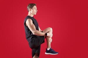 Flexible millennial man stretching his leg over red studio background, side view. Athletic young guy in sportswear warming up before training, practicing flexibility exercises photo