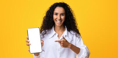 Joyful Brunette Lady Pointing At Smartphone With Black Blank Screen, Showing New App, Isolated On Yellow Background, Mockup Image With Free Space For Your Design, Creative Template For Advertisement photo