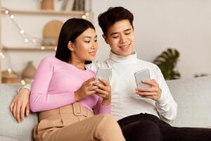 Asian Couple Using Smartphone Phones Browsing Internet Sitting Together On Sofa At Home. Spouses Using Social Media Networking And Texting Online. Mobile Application For Cellphone Concept photo