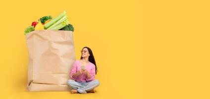 A woman sits on the floor beside a large paper bag filled with colorful vegetables and fruits, smiling as she holds a smartphone. This reflects a shift in shopping practices. photo