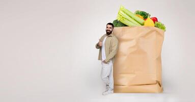 A man stands beside a large paper bag filled with fresh vegetables and groceries. He appears satisfied, reflecting the new normal of shopping during isolated times. photo