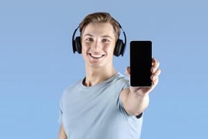 Positive young athlete in headphones showing cellphone with blank screen over violet studio background, mockup for design. Fit sportsman listening to music and holding phone with space for mobile app photo