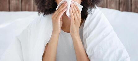 Flu and colds, illness at home and quarantine. Upset young african american lady sits on bed in blanket and wipes nose with napkin, suffering from fever in cozy bedroom interior, free space, cropped photo