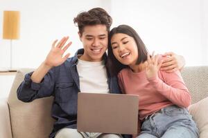Chinese Couple Making Online Call Via Laptop Computer Sitting On Sofa At Home. Spouses Calling Family Members Waving Hello To Screen. Distant Communication Concept photo