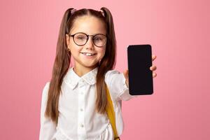 Technology and school. Little child in glasses in uniform showing phone with blank screen to camera, isolated on pink background photo