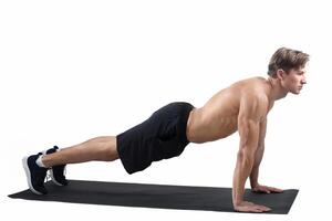 Full length portrait of strong young man standing in plank over light studio background, empty space. Side view of shirtless millennial man doing core exercises on yoga mat photo