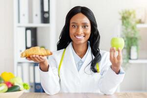 Making healthy food choices. Beautiful female nutritionist holding croissant and apple at table in medical office. African American dietitian selecting between healthy and junk products photo