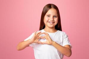 Expression of emotion of love and kindness. Smiling little girl showing heart sign with hands, isolated on pink background, studio shoot photo