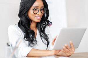Working online concept. Positive millennial black lady using tablet computer at modern office. African American manager having online business call, looking through document on web photo