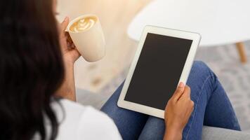 Young black woman with coffee sitting on sofa and using tablet computer with blank screen at home, mockup for your website design. African American lady working online, selective focus photo