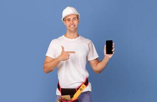 Happy couple in protective helmets pointing at smartphones with black blank screen, recommending home remodeling app or construction materials website, standing over blue studio background, mockup photo
