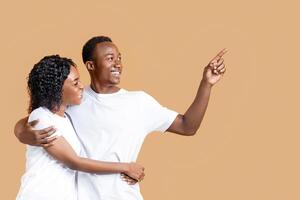 Cheerful black guy showing his girlfriend copy space on yellow studio background. African american lovers hugging and looking at empty space for your advertisement or text, panorama photo