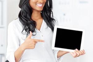 Smiling black business lady pointing at tablet computer with blank screen at office, mockup for app or website design. Successful female manager with touch pad, space for your ad photo
