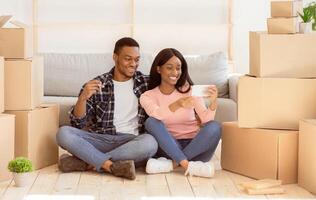 Young black family chatting online with friends, showing key to their new house on camera, sitting among carton boxes on moving day. African American couple having call during relocation photo