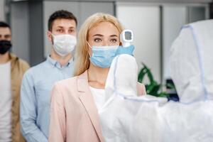 Health control at work and new normal for company. Man in protective suit checks temperature with infrared thermometer at multiracial workers in protective masks keeping social distance, panorama photo