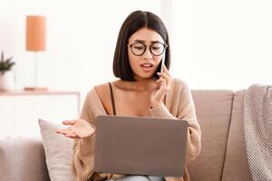 Problem Concept. Frustrated serious woman in glasses talking on mobile phone, sitting on couch and using laptop, having problem with internet connection, calling customer support hotline photo