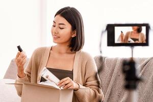 Product Review. Satisfied asian young woman vlogger holding foundation and brush, unpacking cardboard beauty box with cosmetics products, recording for her social media blog on camera at home photo