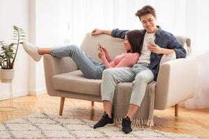 Chinese Young Couple Using Phones Browsing Internet And Texting Relaxing On Weekend Sitting On Sofa At Home. Mobile App For Cellphone, Modern Gadget Lifestyle Concept photo
