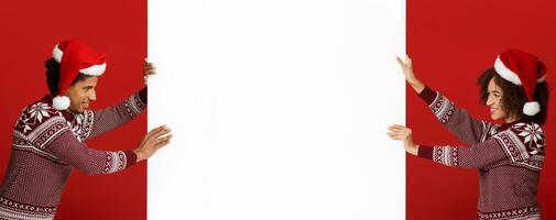 Winter, confrontation and Xmas mood. Cheerful young african american couple in Xmas hats and sweaters, push white billboard with empty space for advert isolated on red background, panorama, side view photo