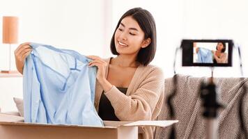 Portrait of happy asian female seller showing stylish blouse on camera on tripod, recording on camera device at home, female vlogger influencer sitting with cardboard box, selling clothes photo