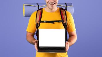 Book your tour online. Positive millennial traveler with backpack showing laptop computer with empty screen on blue studio background. Mockup for tourist agency website photo
