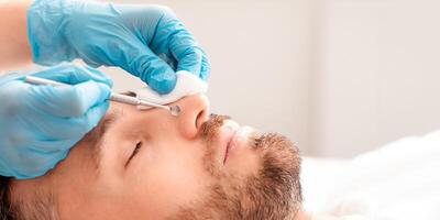 Unrecognizable cosmetologist in medical gloves removing blackheads from middle aged man nose, using tools and cotton pad, side view. Closeup of handsome man attending cosmetologist photo