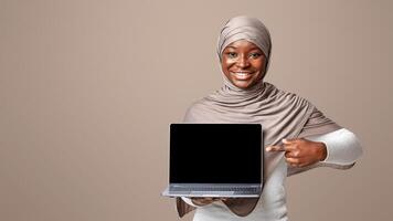 Mockup Image Of African Lady In Hijab Holding Laptop Computer And Pointing At Black Screen, Showing Free Copy Space For Your Website Design, Smiling Muslim Woman Standing Over Light Studio Background photo