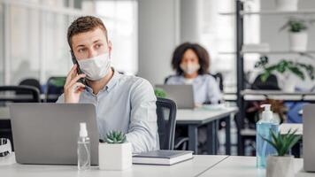 New rules for taking care of health at work after quarantine. Young man in protective mask talking on smartphone with client, at table with laptop and antiseptic in interior of office, free space photo