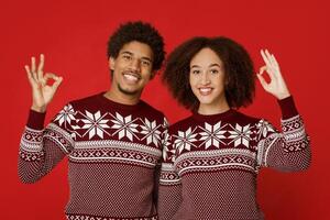 Great New Year and Christmas party. Joyful african american family in identical sweaters show sign okay with their hands and look at camera, isolated on red background, empty space, studio shot photo